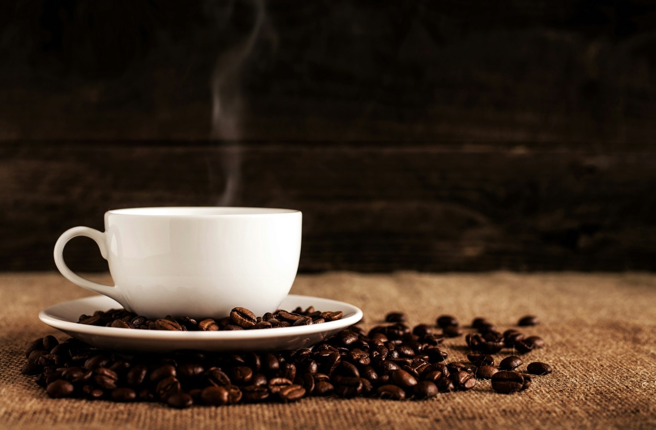 Weißer Keramikbecher mit Untertasse und Kaffeebohnen auf braunem Textil.